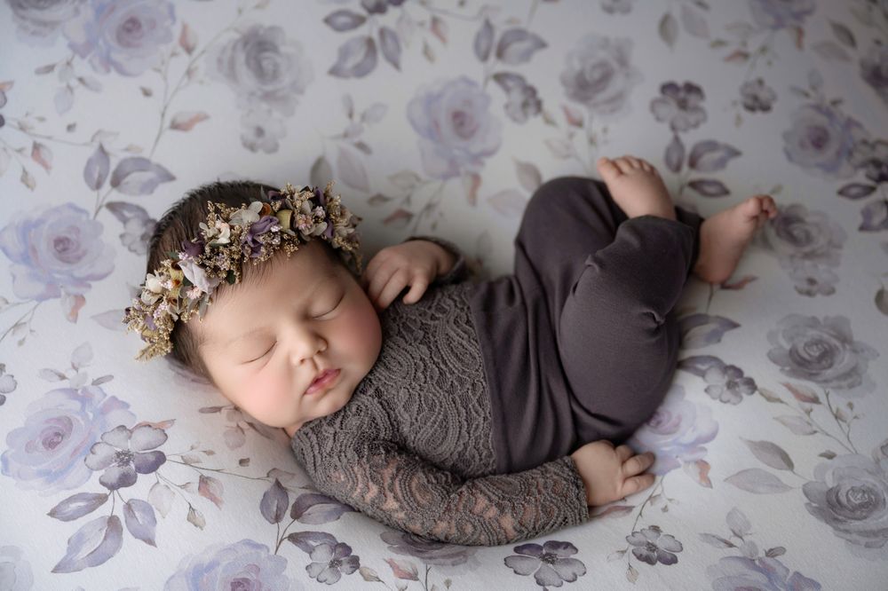 Sleeping newborn in gray lace romper with floral headband on purple rose-print blanket.