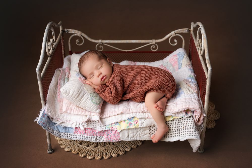 Sleeping newborn in brown knit wrap posed on miniature vintage iron bed with quilts.