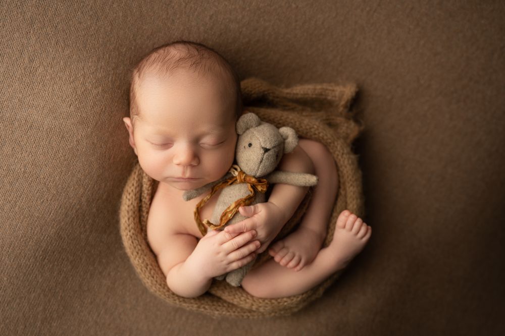 Sleeping newborn baby cuddling small teddy bear in brown knit bowl on brown backdrop.