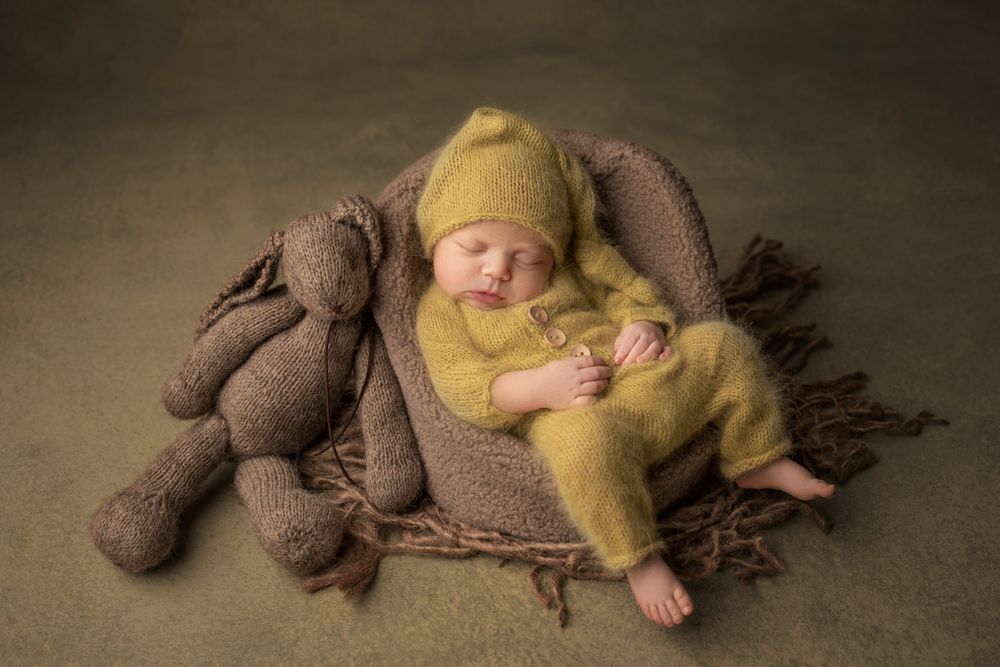 Newborn baby in yellow knit outfit and hat sleeping beside matching stuffed teddy bear.