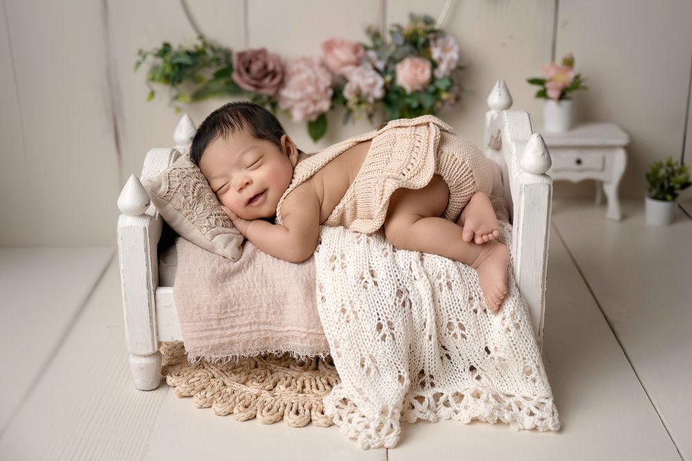 Smiling newborn baby posed on tiny white bed with knit blanket and floral decor.