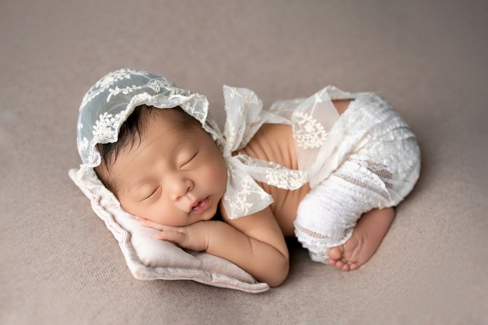 Peaceful newborn baby in a white lace bonnet and romper, sleeping on a small pillow against a gray background.