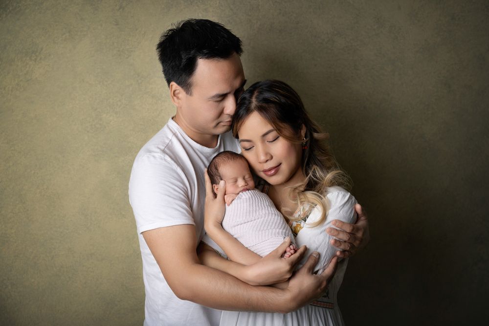 Asian parents in white clothing tenderly holding their sleeping newborn baby together.