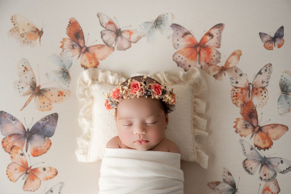 Sleeping newborn wrapped in white swaddle with floral crown surrounded by butterfly decorations.