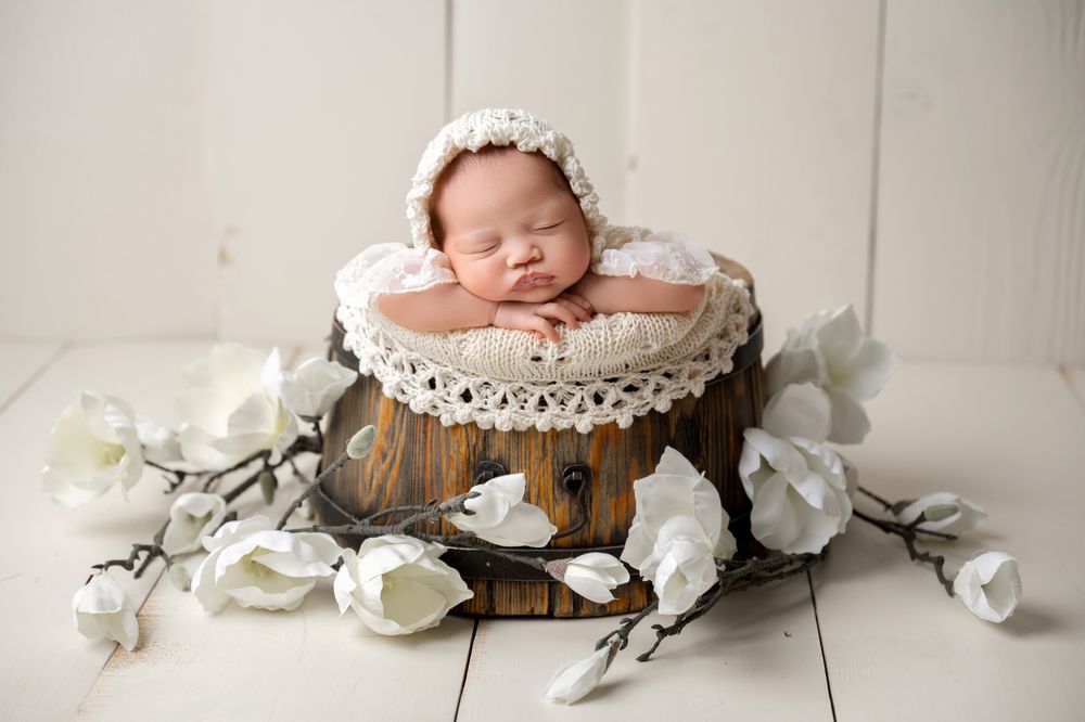 Sleeping newborn baby in white lace-trimmed wooden bucket with white flowers.