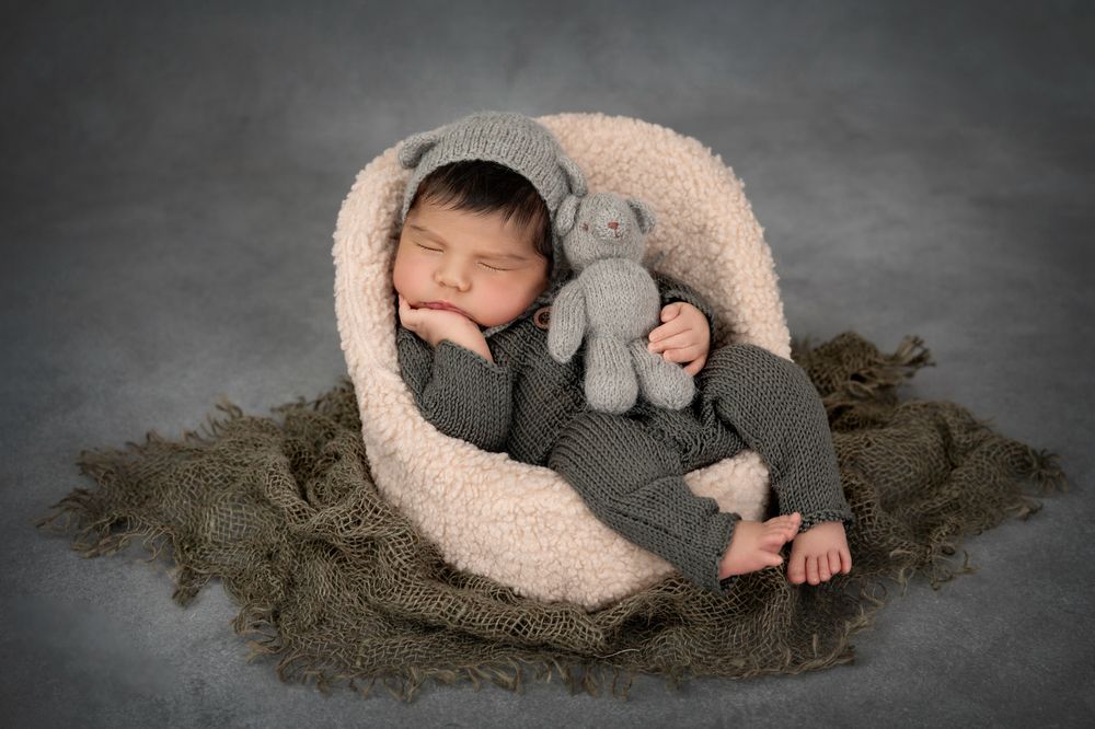 Newborn baby in gray knit outfit sleeping in bowl with matching stuffed elephant toy.
