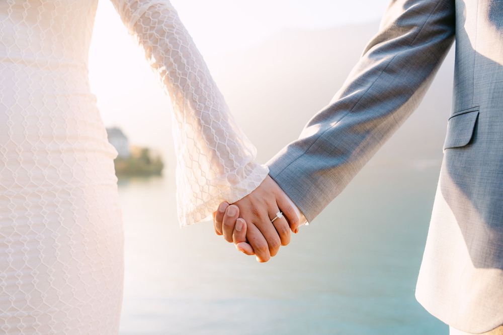 A couple holds hands while wearing wedding attire, showing their rings against a soft blurred background in Iseltwald.