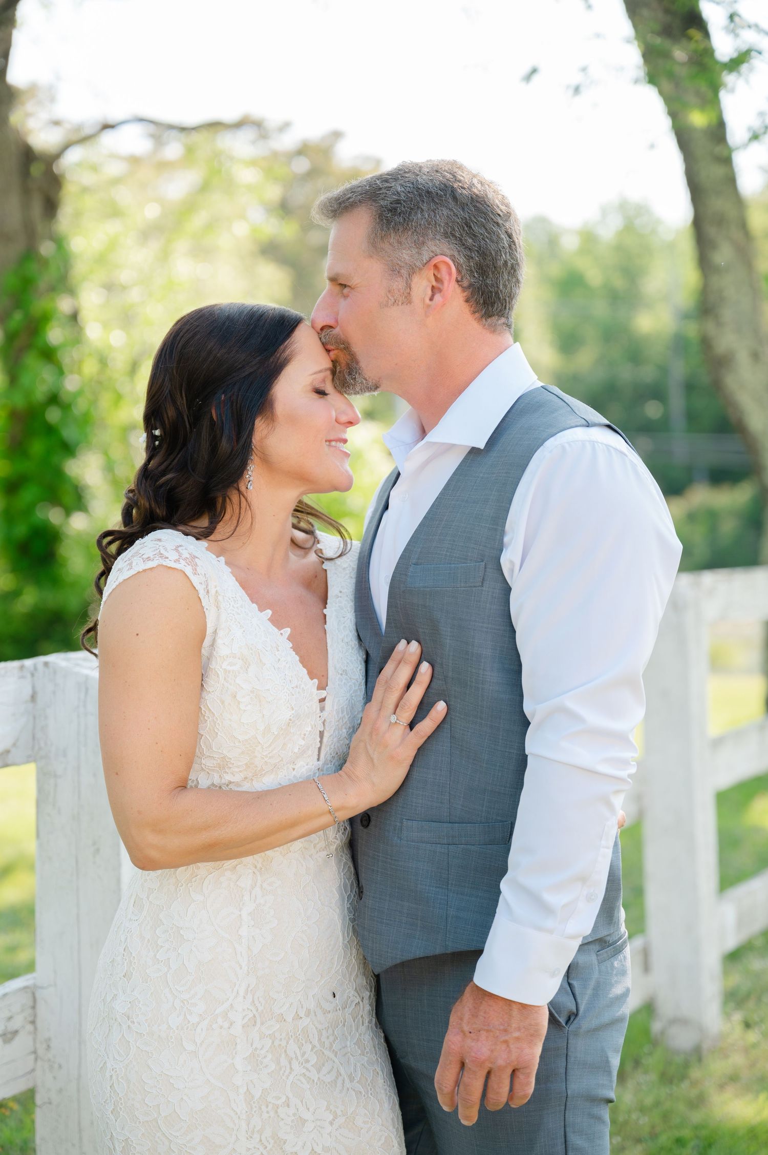 A couple shares an intimate moment outdoors by a white fence, the bride wearing lace and groom in a grey vest.