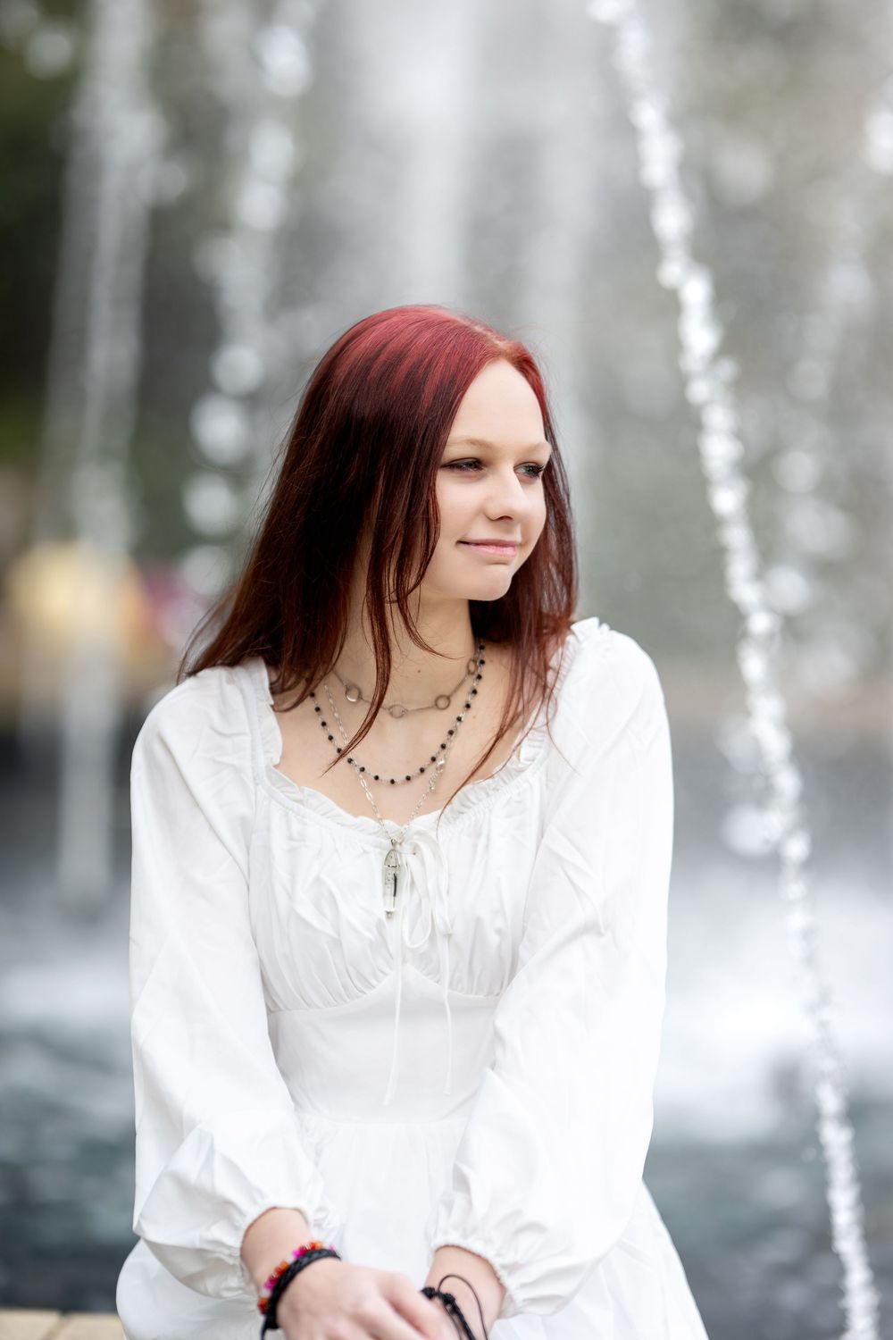 Red-haired young woman in white top with necklaces sits near fountain jets in soft outdoor light.