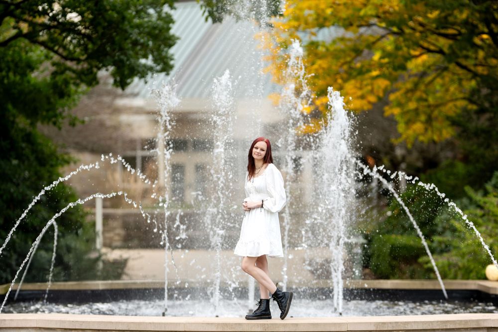 Young woman in white dress poses among arching fountain jets with yellow autumn trees in background.