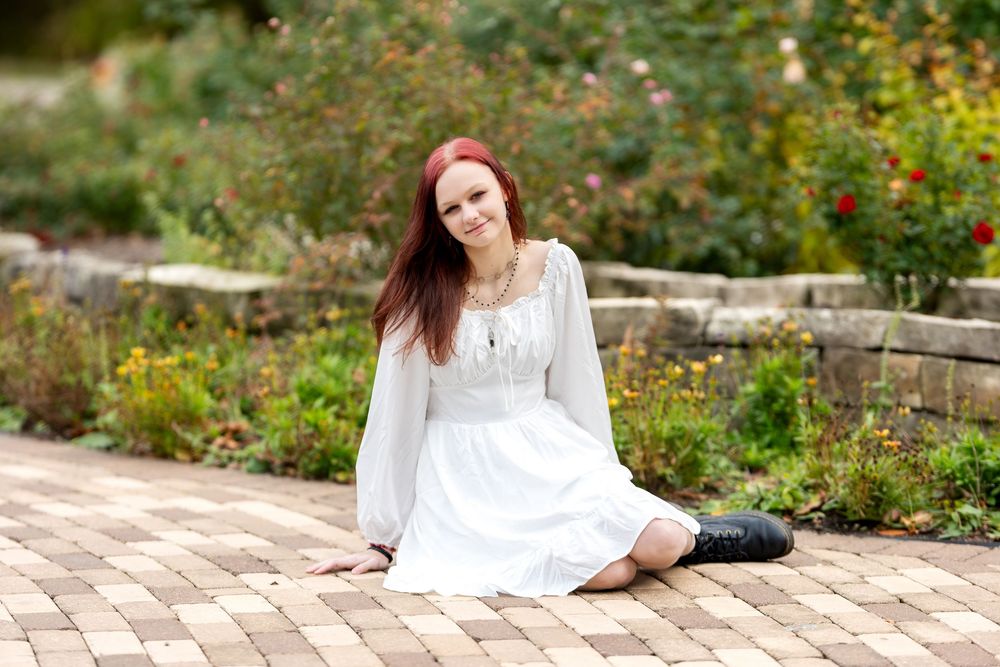 Smiling redhead in white dress sits on brick path in colorful garden with roses in background.