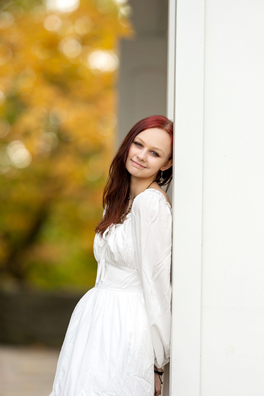 Smiling redhead woman in white dress peeks around white column with vibrant yellow fall foliage background.