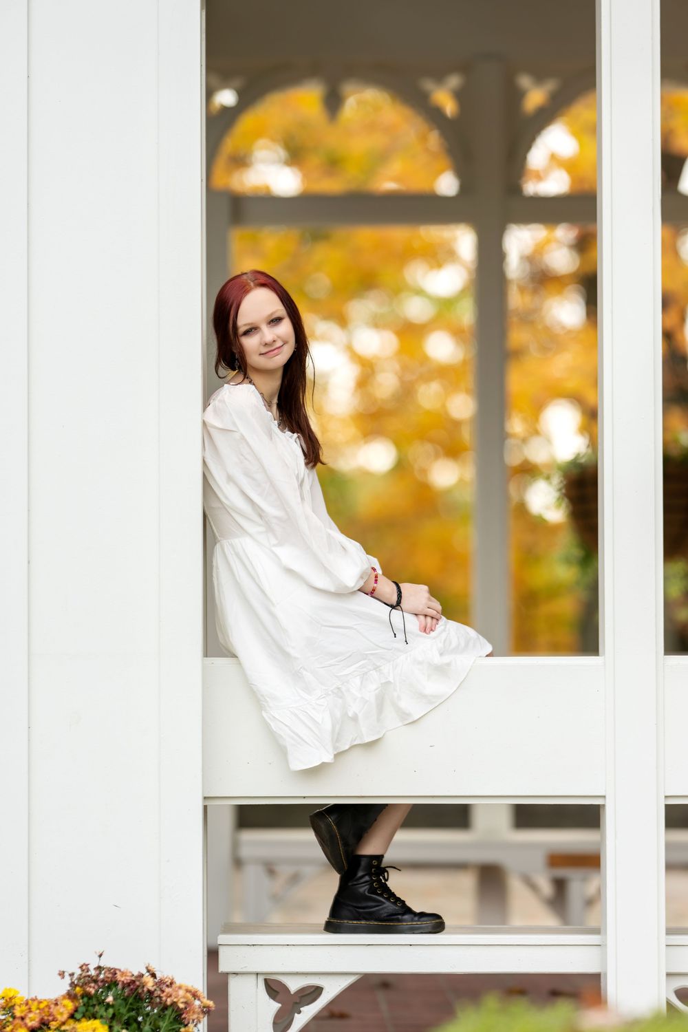 Young redhead woman in white dress and black boots sits on white gazebo railing amid autumn scenery.