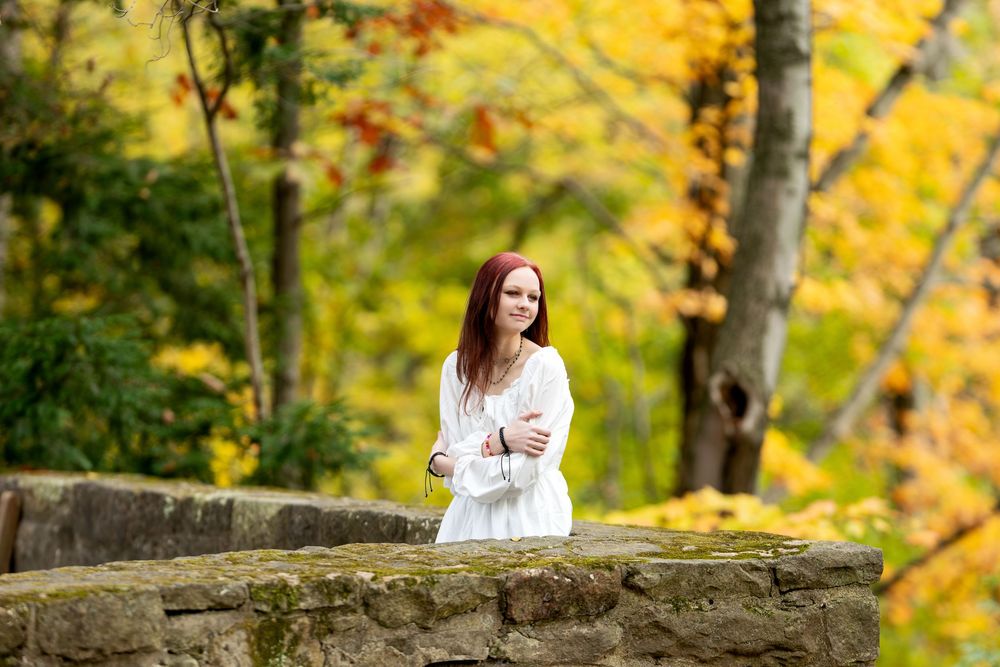 Young redhead woman in white dress with crossed arms stands before stone wall with golden fall trees.