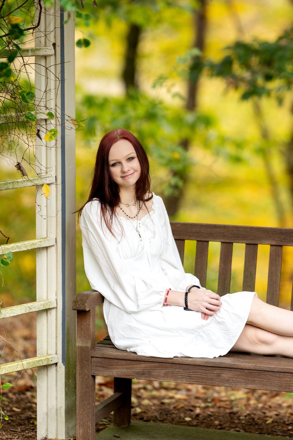 Smiling redhead woman in white dress sitting barefoot on wooden bench in autumn garden gazebo.