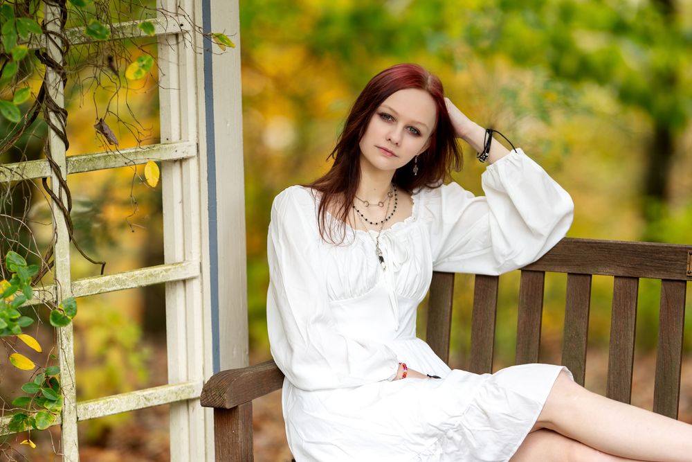 Redhead woman in white dress seated on wooden bench by white lattice with colorful fall leaves.