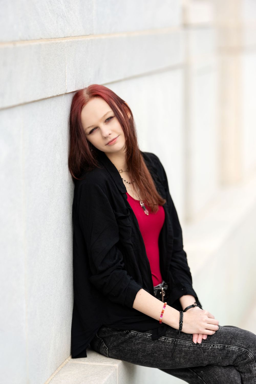 Young redhead woman in black jacket and red top leaning against white marble wall.