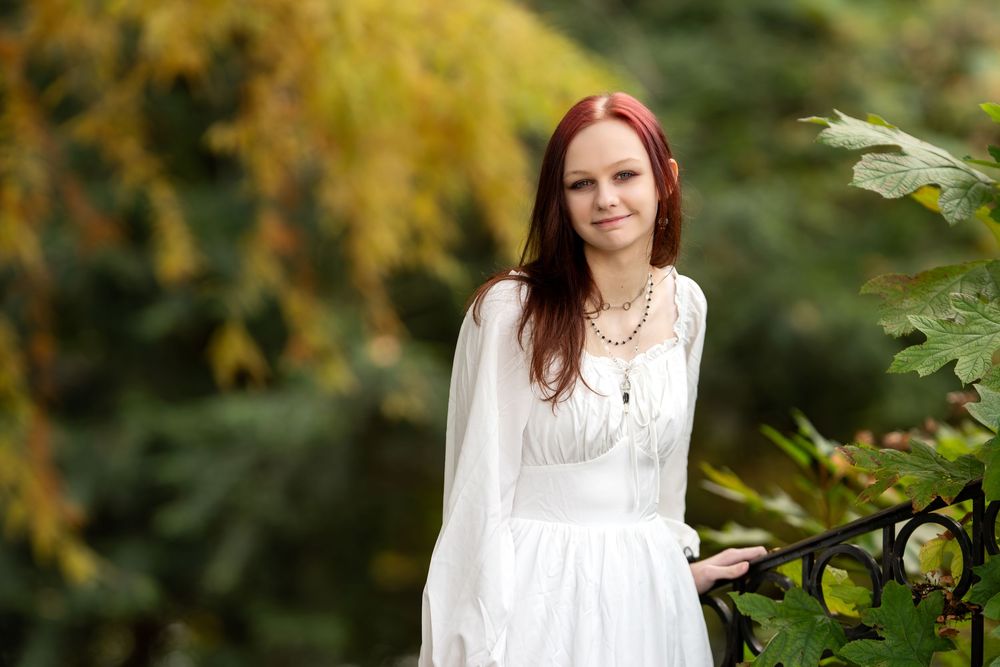 Red-haired woman in white long-sleeve dress smiling near iron fence with colorful fall foliage.