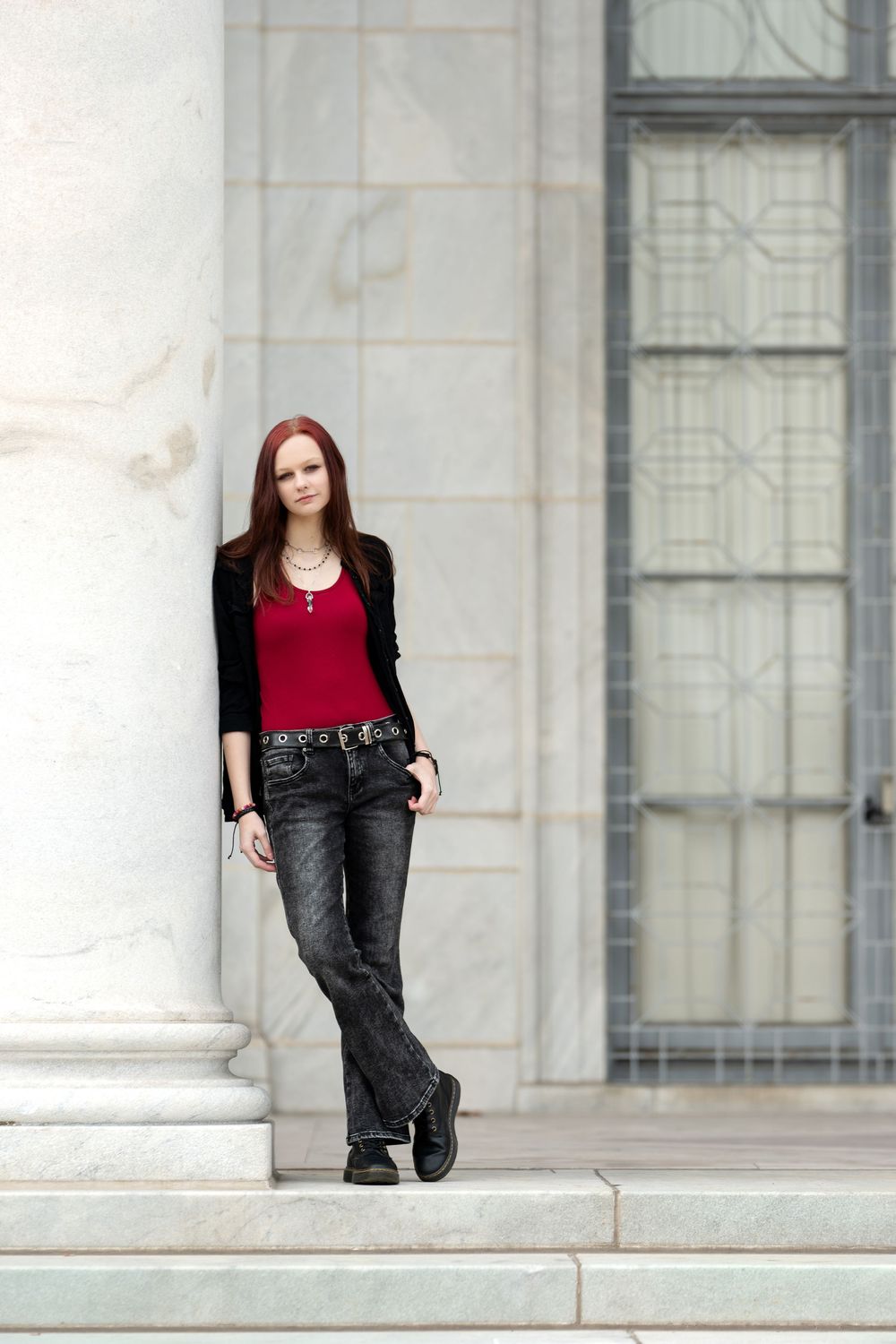 Young redhead woman posing by marble column in black jacket and gray jeans.