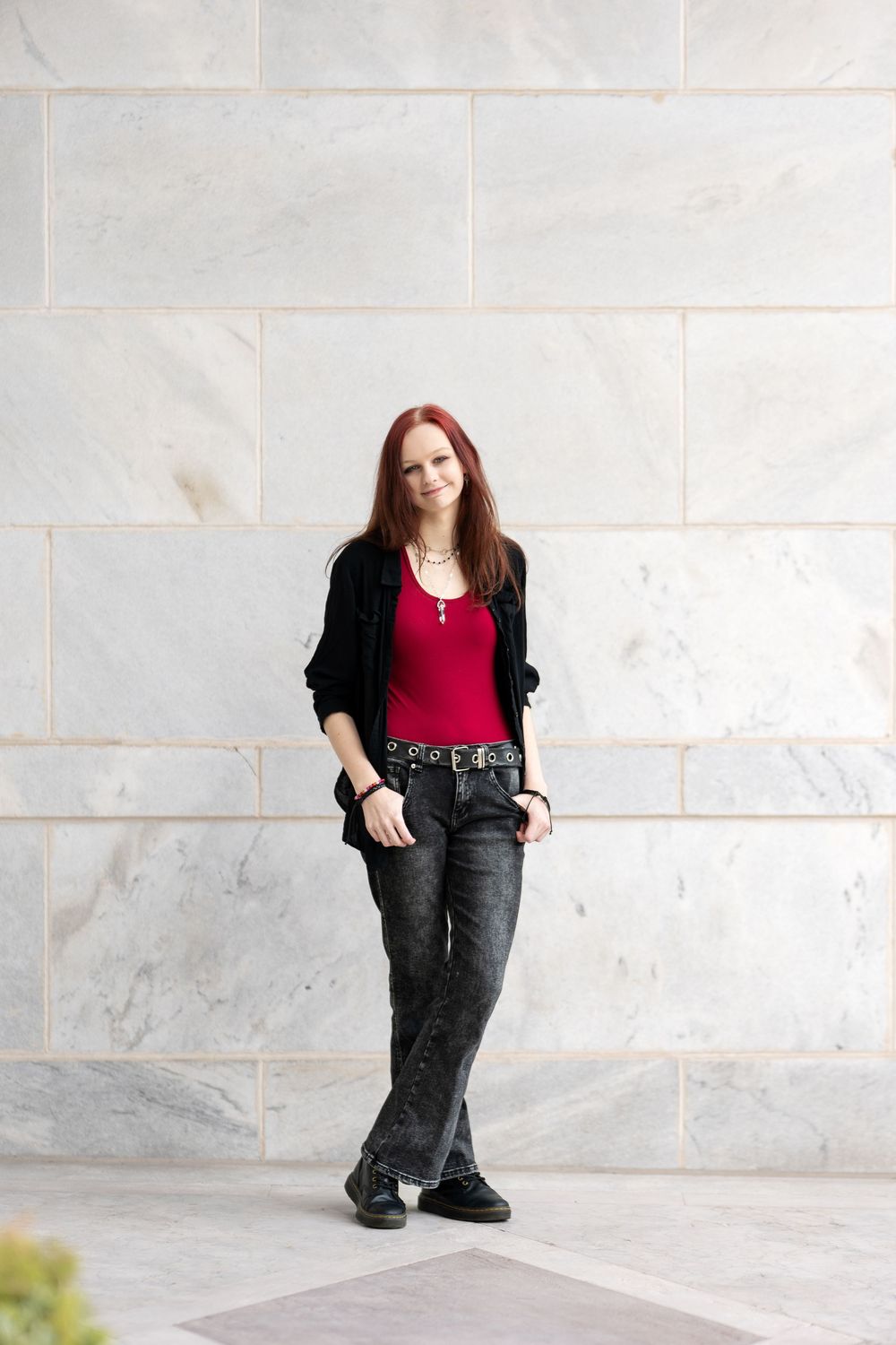 Young woman with red hair in black jacket and red top standing against marble wall.