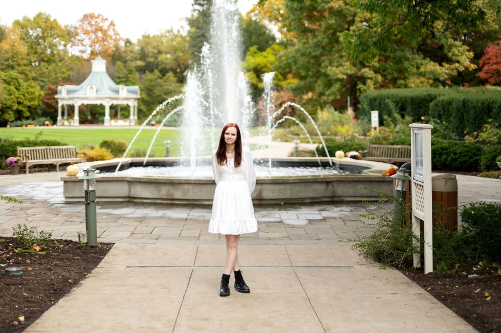 Red-haired woman in white dress standing before large fountain in scenic outdoor park.