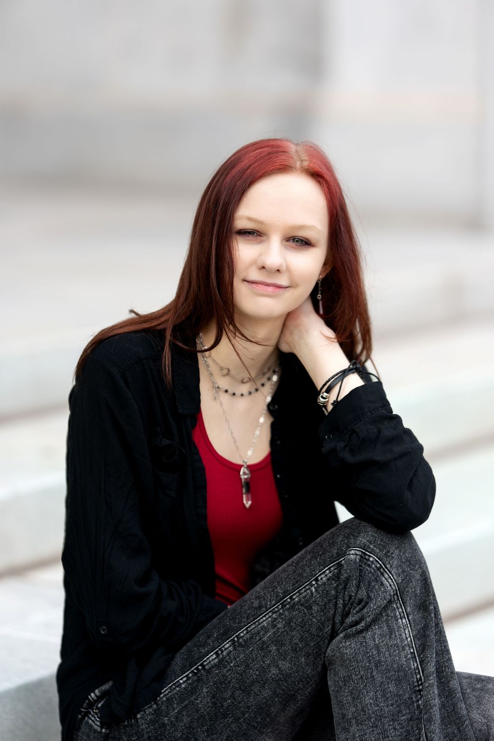 Redhead woman in black jacket and red top sitting on steps outdoors.