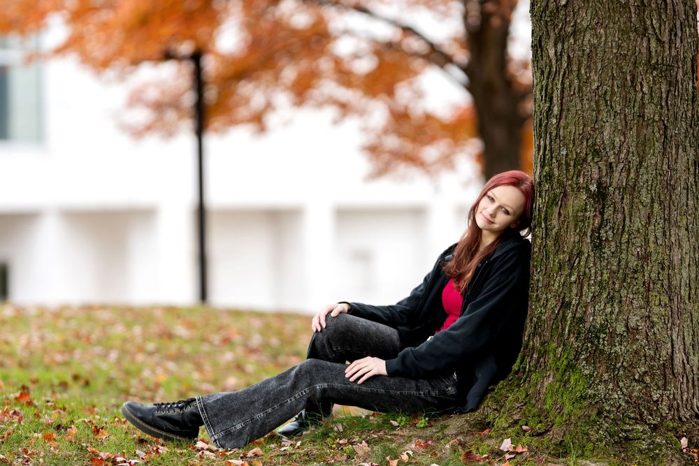 Redhead girl sitting against tree trunk in autumn park with fallen leaves.