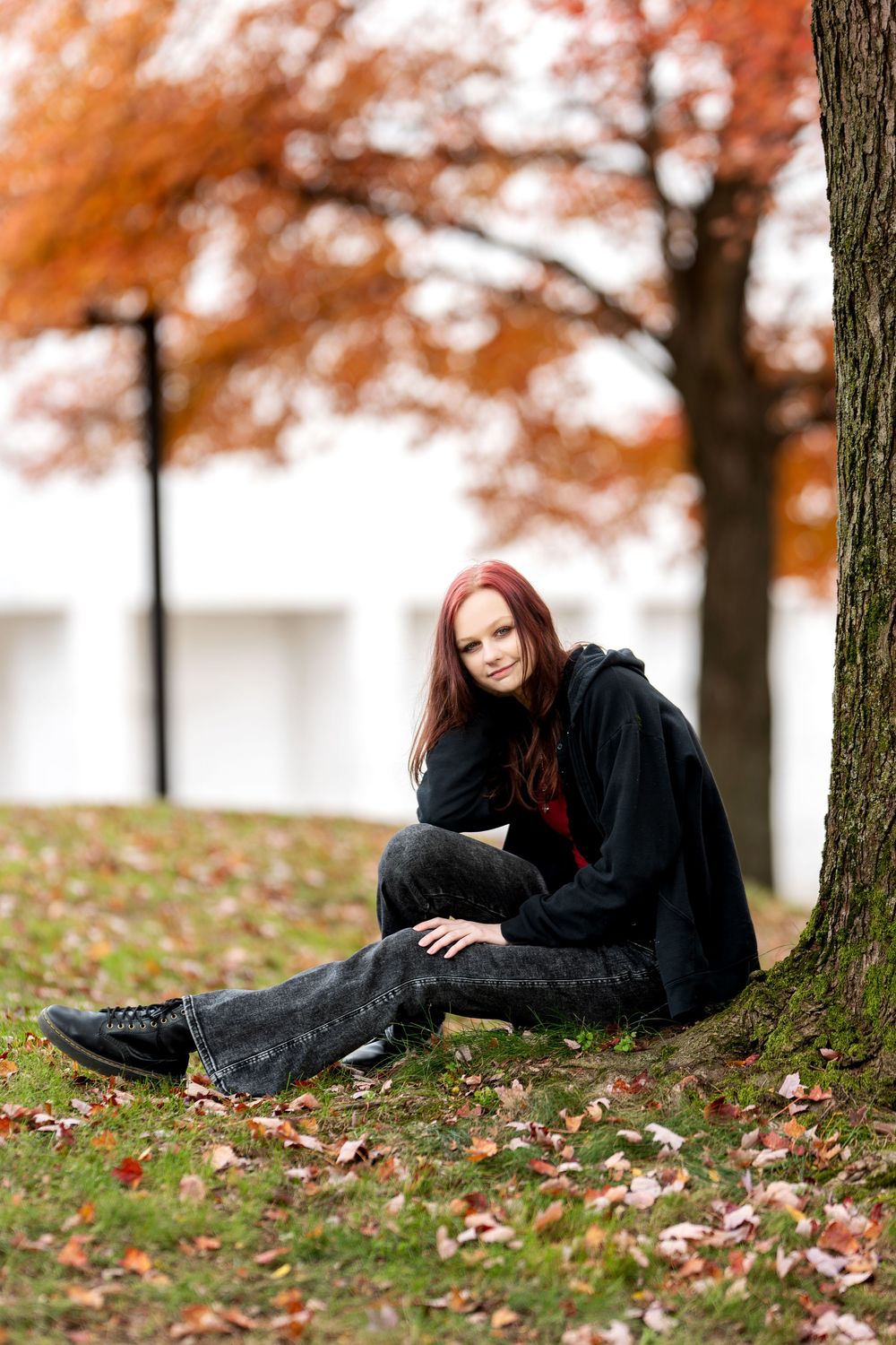 Redhead young woman sitting by tree in autumn park with colorful foliage.