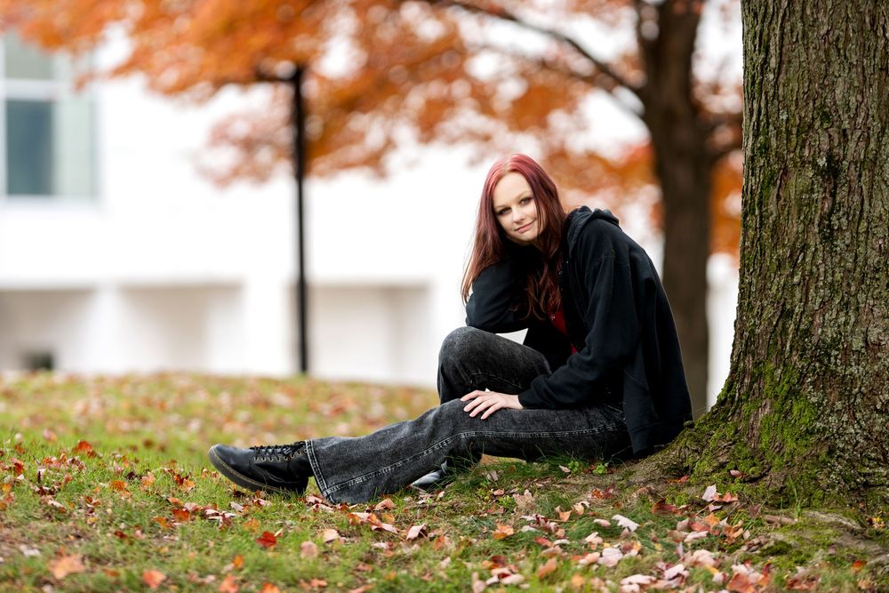 Smiling redhead girl sitting by tree in autumn park with fallen leaves.
