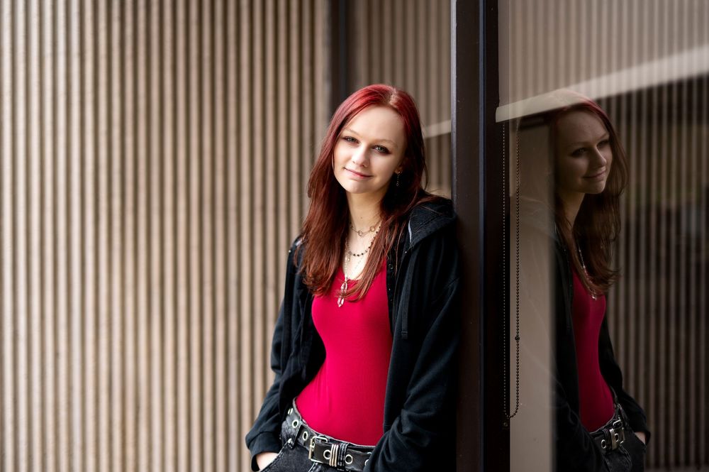 Young woman with red hair in red top and black jacket leaning against striped wall with mirror reflection.
