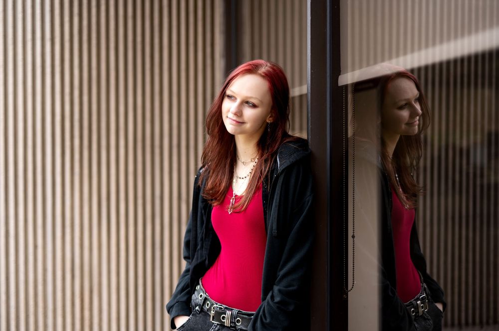 Red-haired woman in red top and black hoodie posing beside glass reflection on striped wall.