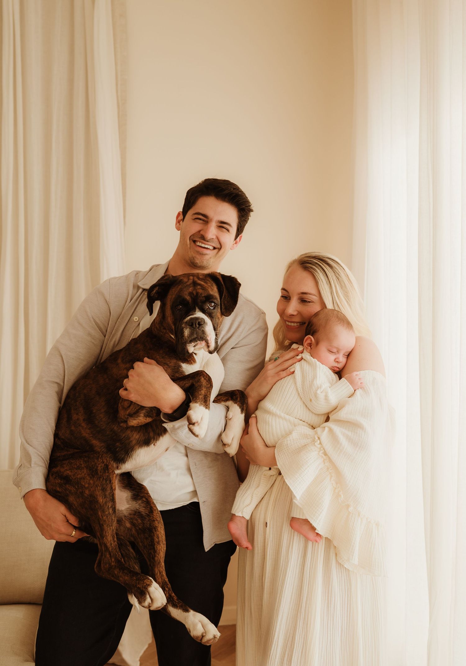 A family stands together indoors near white curtains with their brindle boxer dog and infant wrapped in white.