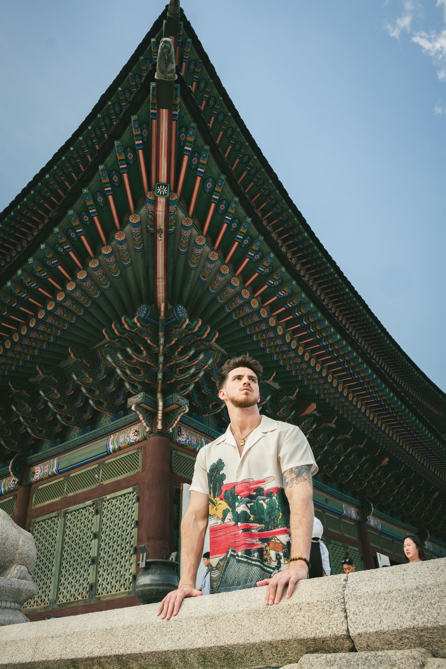 A person sits against the ornate traditional Korean palace architecture with green tiled roofs under blue sky.