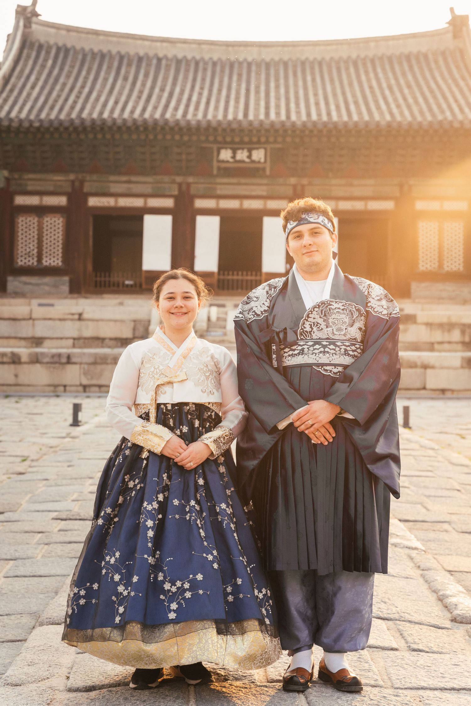 Traditional Korean couple wearing hanbok poses at historic palace courtyard during golden sunset hour.