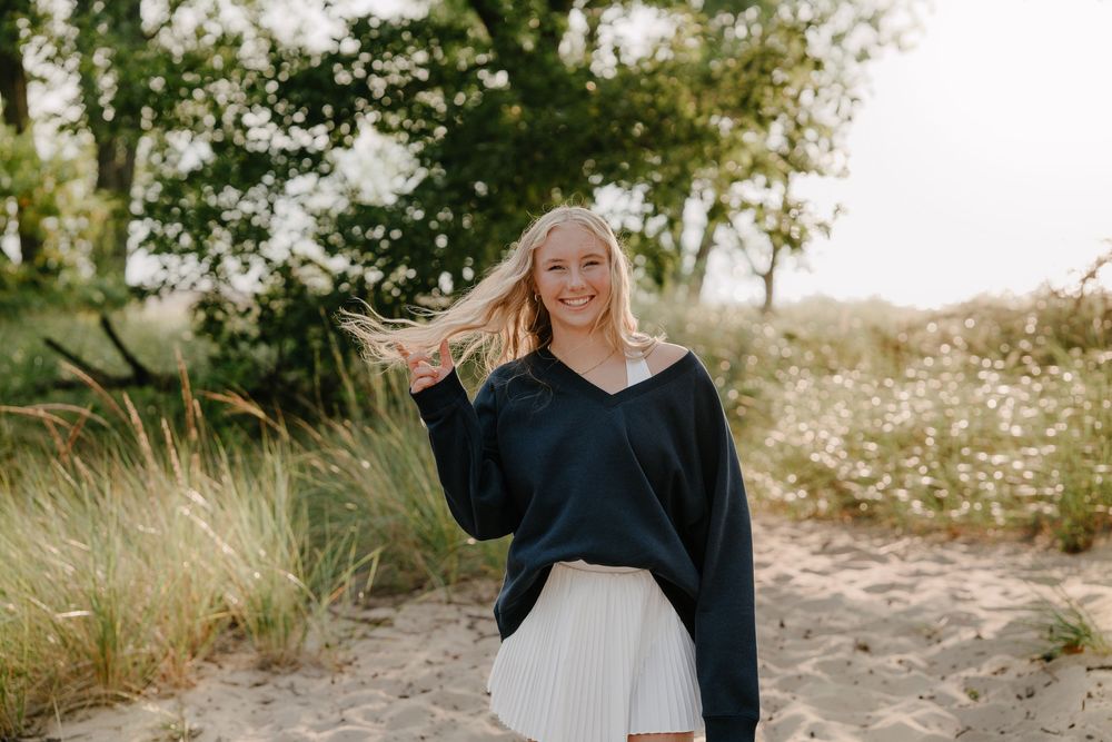 Woman in black sweater poses outdoors in natural sunlit setting with tall grass and trees in background.