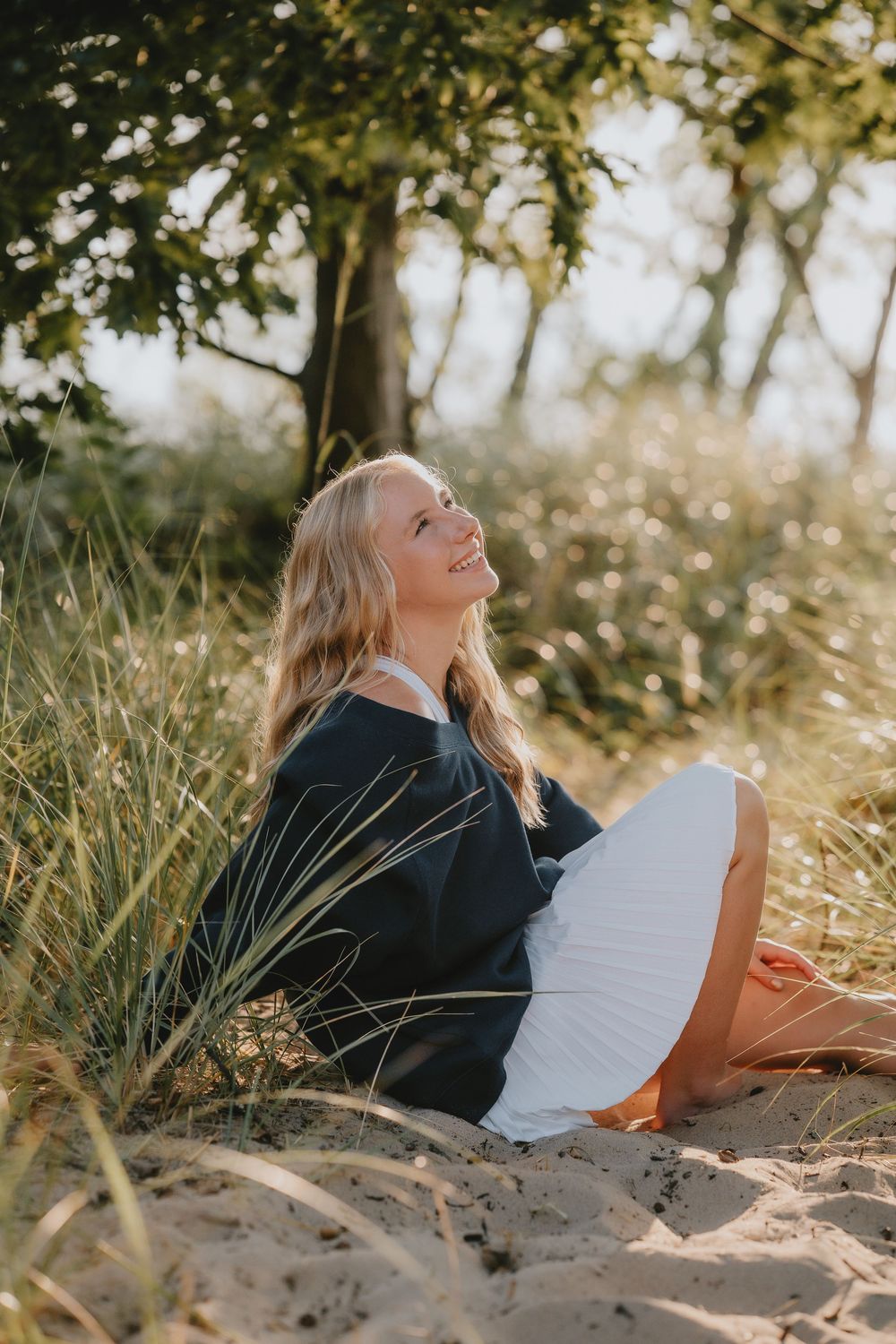 A person in a black sweater sits on sandy beach surrounded by tall grass and dappled sunlight.