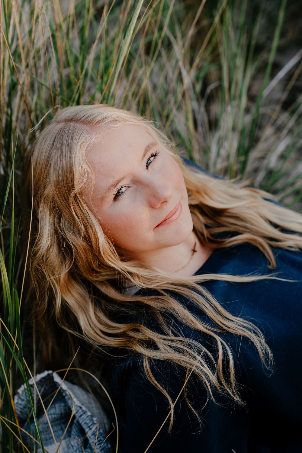 Portrait in tall grass showing blonde hair catching golden sunlight against a dark sweater.