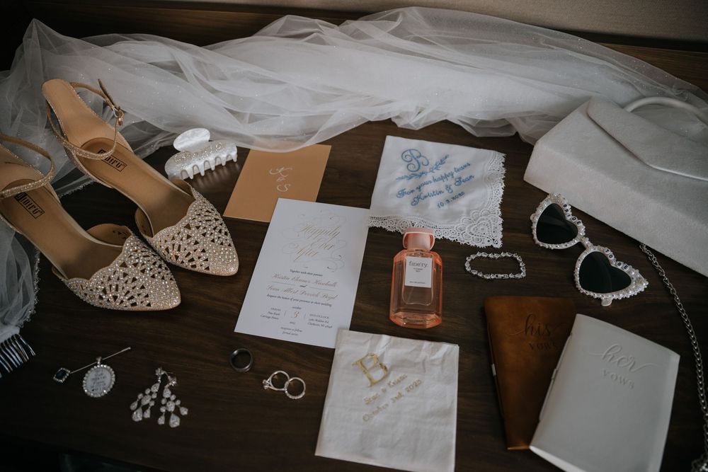 Wedding accessories including perfume, jewelry, shoes, and invitation cards arranged on a dark wooden surface with white tulle.