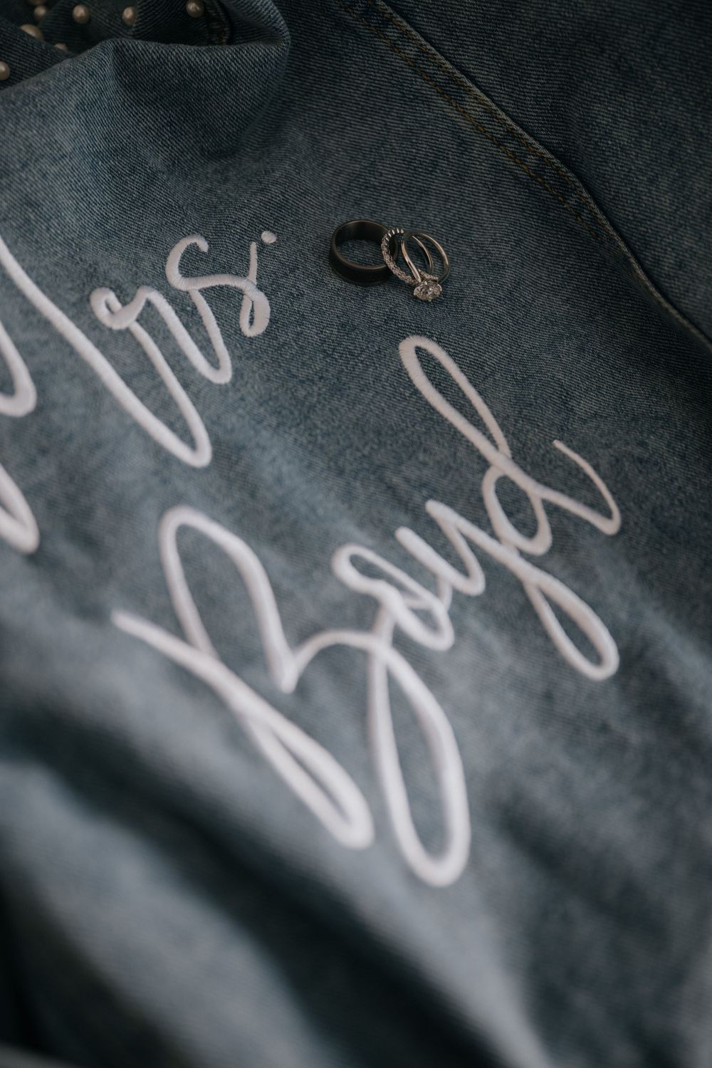 Two wedding rings rest on a dark gray fabric with cursive text embroidered on it.