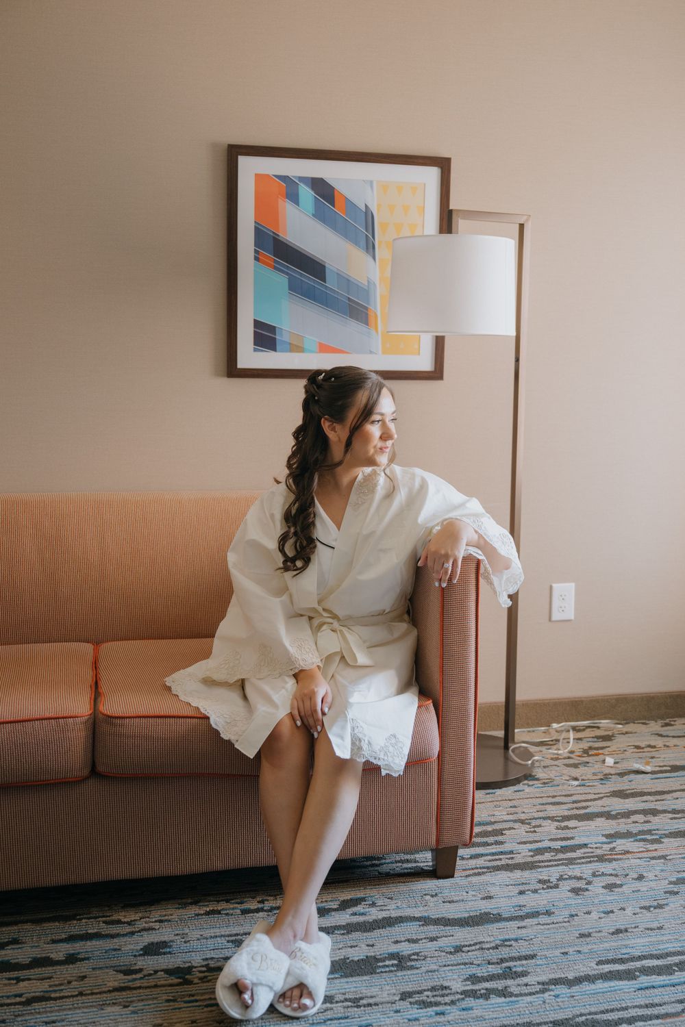 A figure in a white robe sits on a brown couch in a hotel room with modern art decor and patterned carpet.