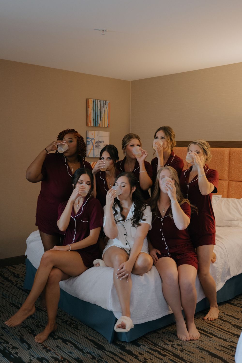 A group of friends sit on a hotel bed taking shots of drinks together in a celebratory moment.