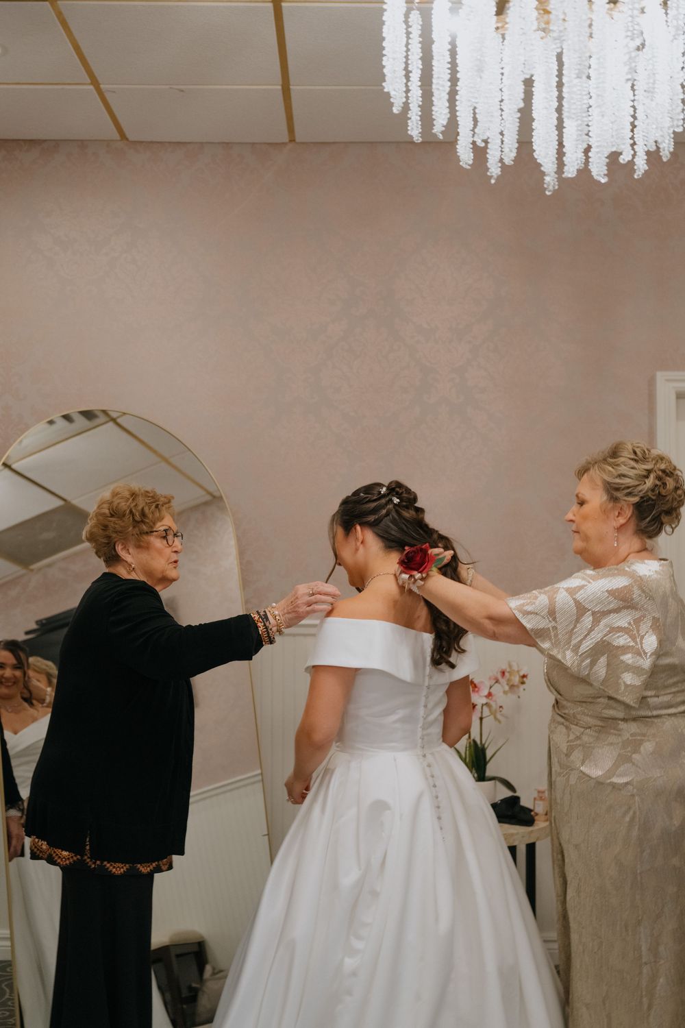 A bride in a classic white off-shoulder gown gets help with final preparations under an elegant crystal chandelier.