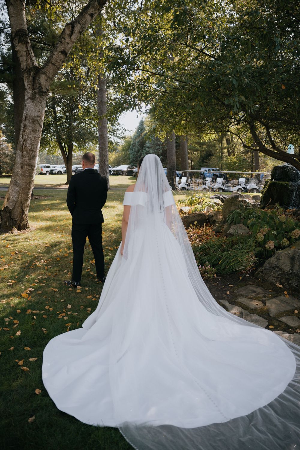 A bride in a flowing white gown with a long train stands behind a suited figure on a lawn surrounded by autumn trees.