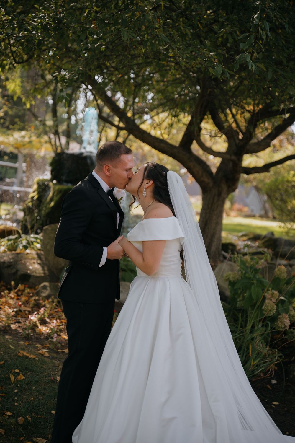 A romantic wedding couple shares a tender kiss under autumn trees in an outdoor garden setting.