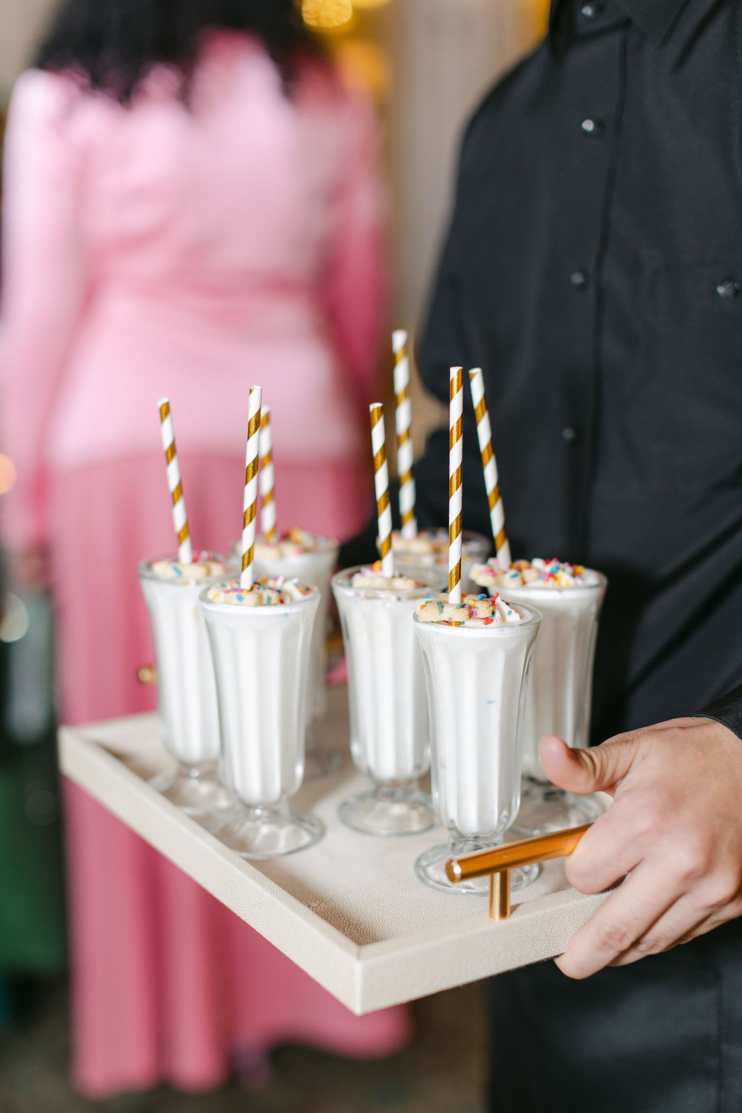 A server carries a white marble tray with miniature milkshakes topped with whipped cream and gold striped straws.