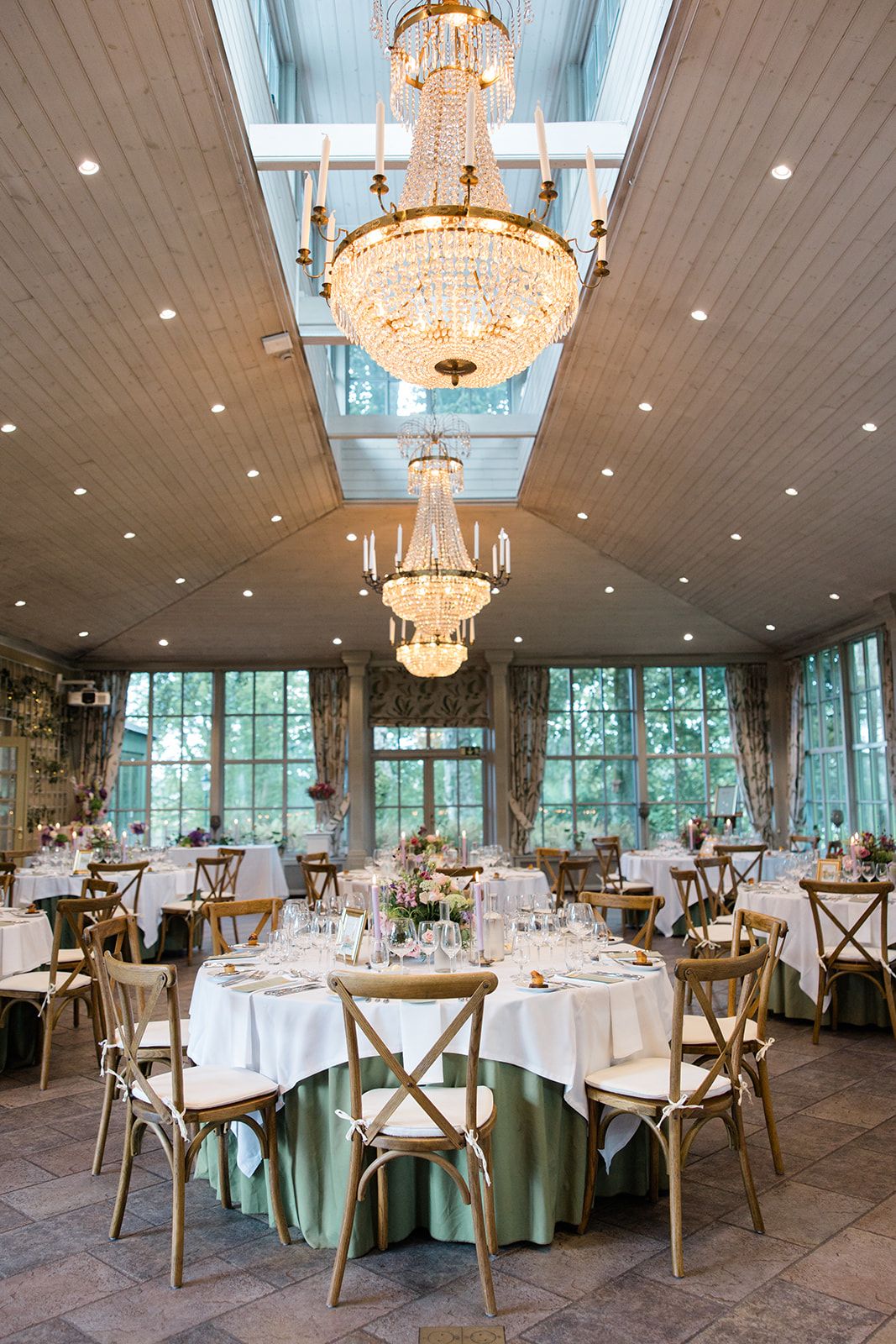 Elegant wedding reception venue with crystal chandeliers, white tablecloths, and wooden crossback chairs in a bright sunlit room.