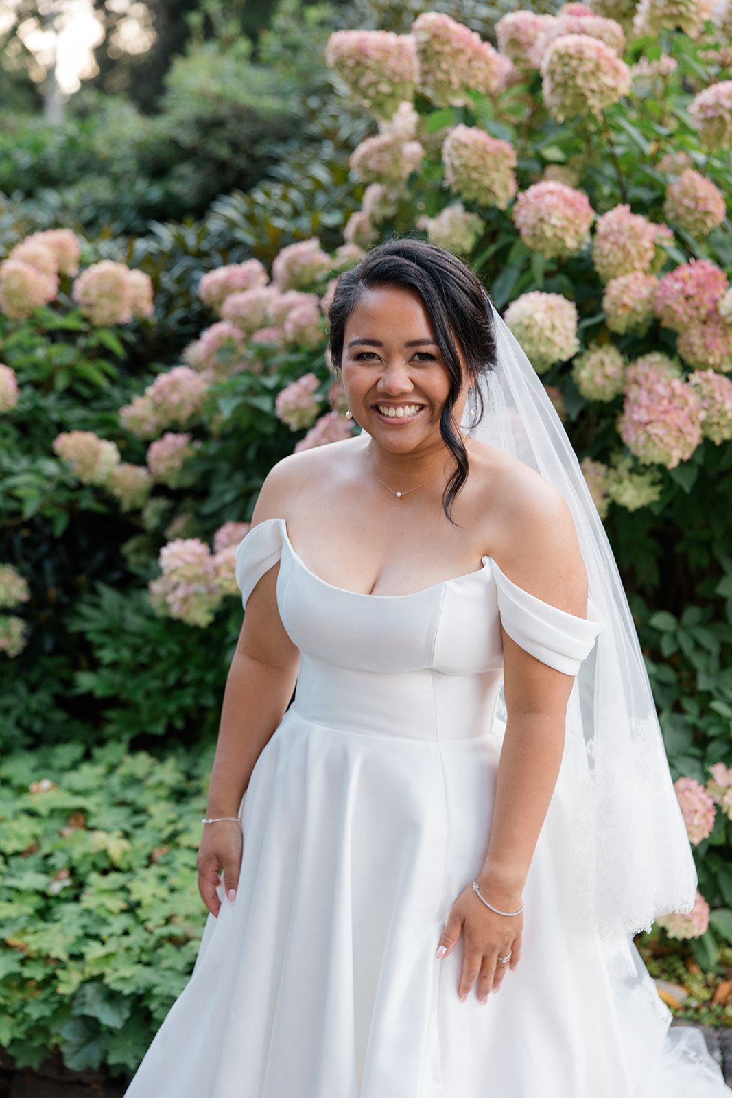 A bride in an elegant white empire waist dress poses in a garden with blooming pink hydrangea flowers.