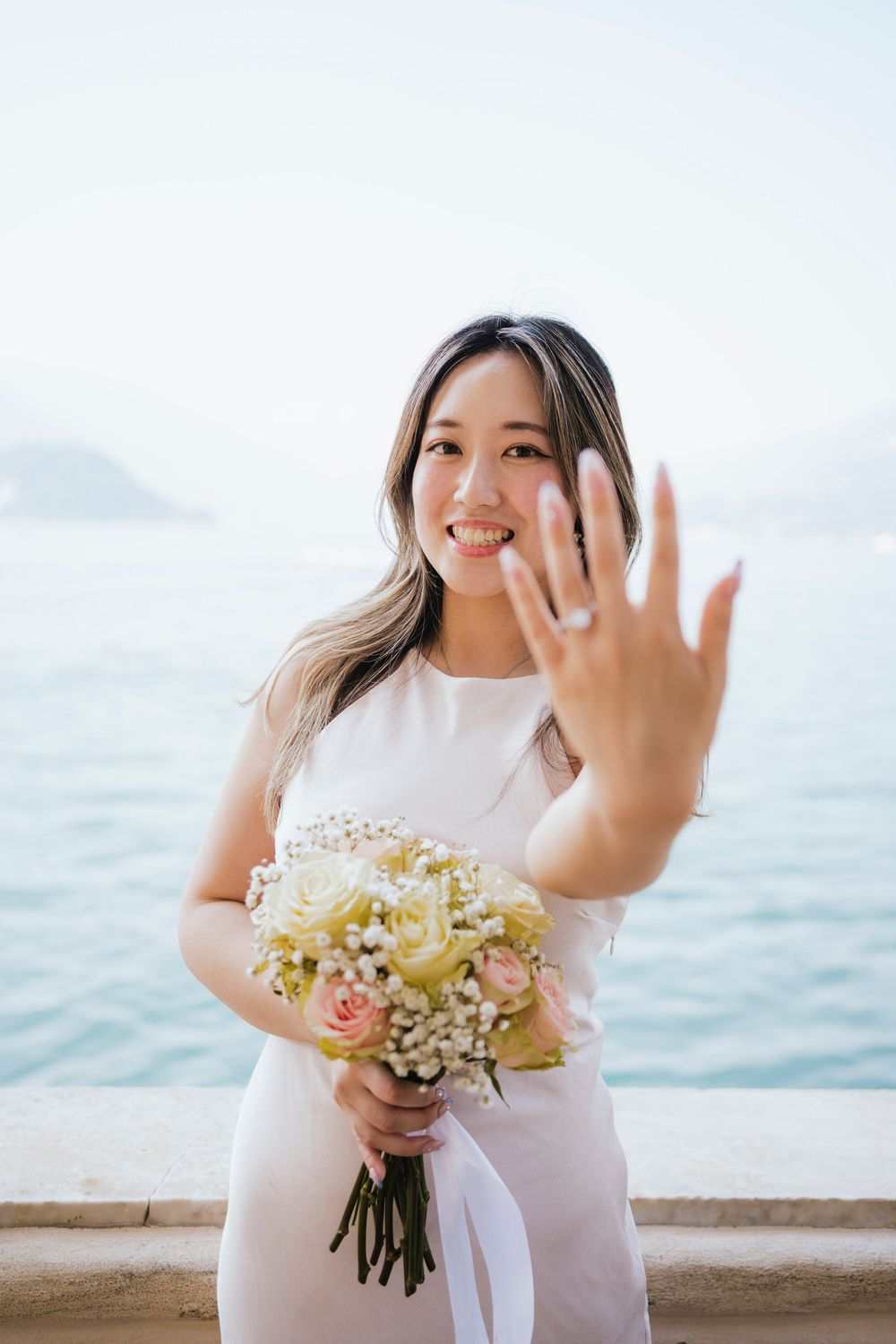 A person in a white dress holds a bouquet of flowers by the ocean, showing off their engagement ring in multiple poses.