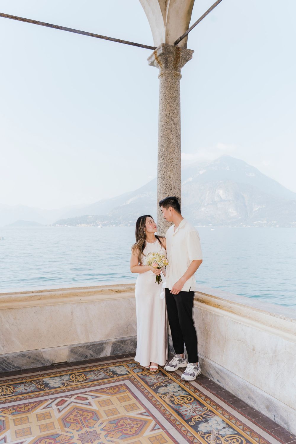 A couple shares a romantic moment on an ornate terrace overlooking a scenic mountain lake in Italy.