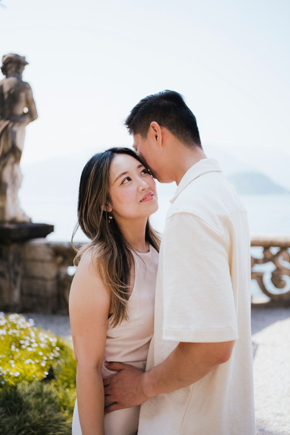 A romantic couple in white outfits sharing intimate moments by a waterfront setting with flowers.